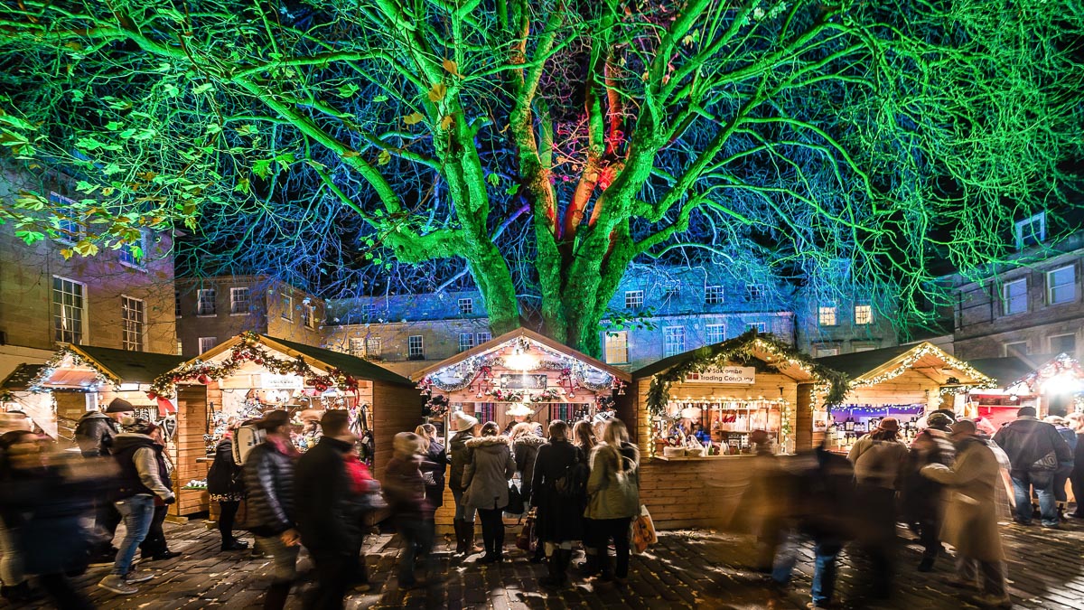 Tree lit up green from Christmas lights behind bath Christmas market stalls 